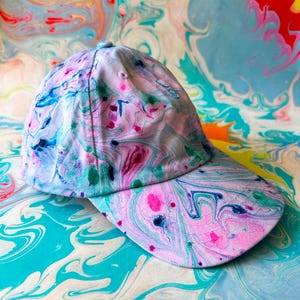 May include: A baseball cap with a vibrant, marbled design in pink, blue, and green hues. The cap features a curved peak and a button detail. The background has a matching marbled pattern.