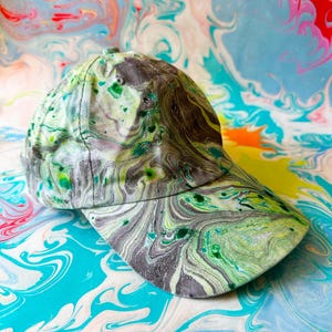 May include: A baseball cap with a marbled pattern in shades of green, grey, and white. The cap has a curved peak and a button on top. The background features a colourful, swirled design in blue, pink, yellow, and white.
