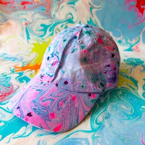 May include: A baseball cap with a swirling, abstract pattern in shades of pink, blue, and purple. The tie-dye effect covers the entire cap, including the brim and crown. This colourful accessory would make a bold fashion statement.