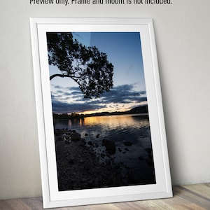 May include: A photograph of a lake at sunset with a silhouetted tree in the foreground. The water is calm and reflects the orange and pink colors of the sky.