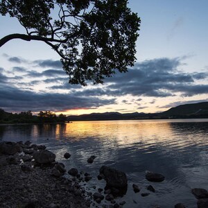 May include: A silhouette of a tree against a cloudy sky with a lake in the foreground. The sky is a mix of blue and orange, with the sun setting in the distance. The lake is calm and reflective, with a few rocks visible in the foreground.