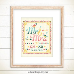 May include: A framed cross-stitch artwork with the words "Mr & Mrs" in teal and pink, adorned with birds, arrows, and hearts. The date "22.09.2015" is displayed below the names. The artwork is set against a cream background.