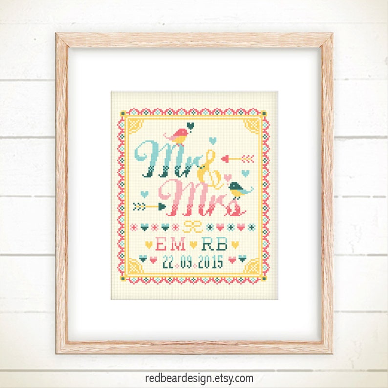 May include: A framed cross-stitch artwork with the words "Mr & Mrs" in teal and pink, adorned with birds, arrows, and hearts. The date "22.09.2015" is displayed below the names. The artwork is set against a cream background.