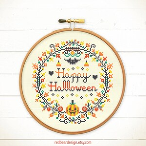 May include: A cross-stitch embroidery hoop with a Halloween theme. The design features the words "Happy Halloween" in black and orange, surrounded by a wreath of colorful autumn leaves, pumpkins, and a bat. The hoop is made of wood.