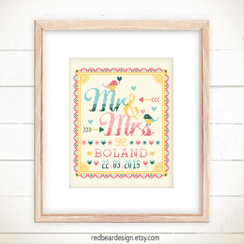 May include: A framed cross-stitch artwork with the words "Mr & Mrs" in teal and pink, along with the name "BOLAND" and the date "22.09.2015". The design includes birds, hearts, and arrows in a folk art style. The frame is light brown.