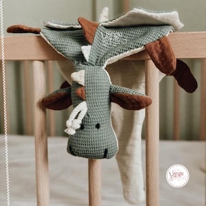 Puede incluir: Un juguete de peluche de dragón de tejido de gofre verde y marrón con una cola blanca y marrón. El dragón está colgado de una barandilla de cuna de madera. El juguete está etiquetado como "Lange Design".