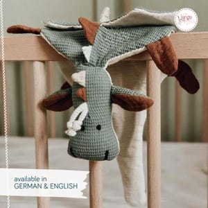 May include: A sage green waffle-textured dragon security blanket featuring brown fabric accents on the wings and head, a white rope detail, and a cream-coloured underside. The dragon has embroidered black eyes and a stitched mouth on its head.