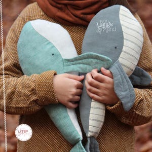 以下が含まれることがあります： 茶色のセーターを着た人が、ターコイズブルーとグレーの2つのぬいぐるみクジラのおもちゃを持っています。グレーのクジラには白いストライプと「von Lange Hand」という刺繍が施されています。