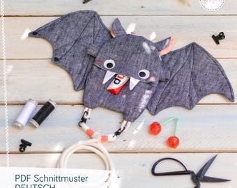Può includere: Una pochette in tessuto grigio a forma di pipistrello con zanne bianche, occhi rotondi e orecchie rosa. La borsa presenta un manico in corda bianca, clip nere e una barretta di cioccolato in bocca. Testo: PDF Schnittmuster DEUTSCH.