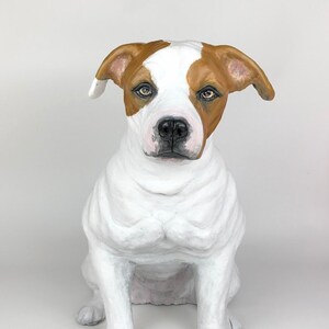 May include: A white and brown dog sculpture, sitting with its front paws together. The dog has a white body with brown patches on its head and ears. The sculpture is made of a material that looks like paper mache.