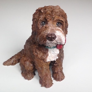 Könnte beinhalten: Eine braun-weiße Hundefigur mit strukturierter Oberfläche. Der Hund hat ein rotes Halsband mit grünem Anhänger und sitzt mit leicht herausgestreckter Zunge. Die Figur steht auf einer weißen Oberfläche.