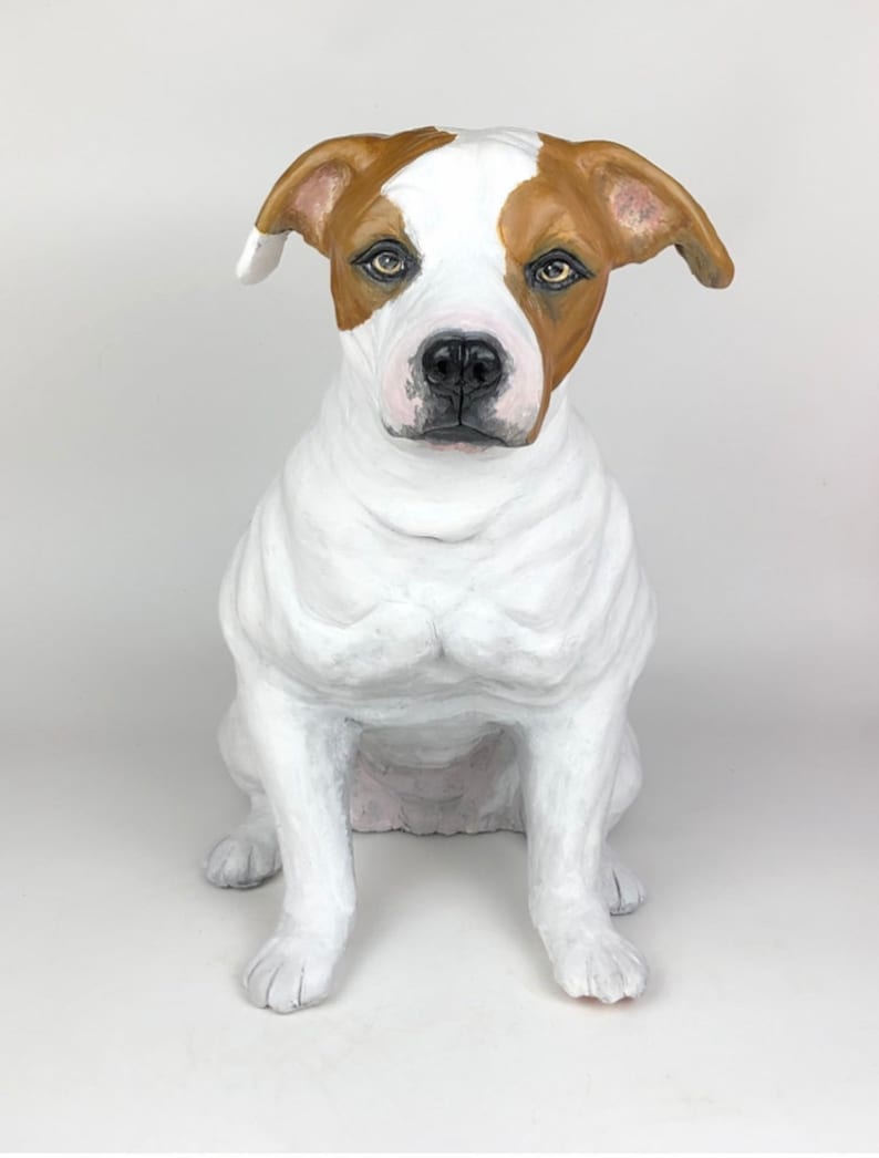May include: A white and brown dog sculpture, sitting with its front paws together. The dog has a white body with brown patches on its head and ears. The sculpture is made of a material that looks like paper mache.