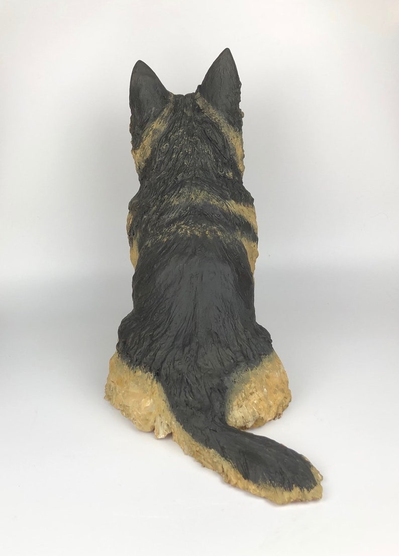 May include: A black and tan German Shepherd puppy figurine sitting with its tail curled behind it. The figurine is made of a textured material that resembles fur.