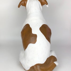 May include: A white and brown ceramic dog sculpture sitting on a white surface. The dog is facing away from the camera with its tail curled around its legs.