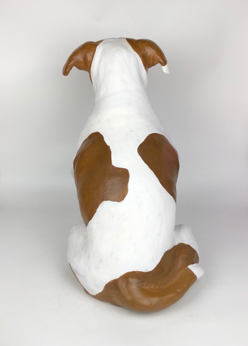 May include: A white and brown ceramic dog sculpture sitting on a white surface. The dog is facing away from the camera with its tail curled around its legs.