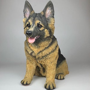 May include: A ceramic figurine of a German Shepherd dog sitting with its tongue sticking out. The dog is brown and black with a white patch on its chest.
