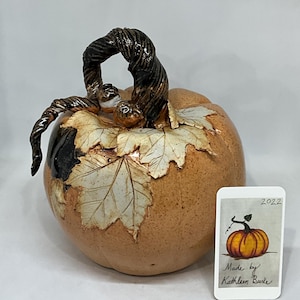 May include: A ceramic pumpkin with a brown stem and leaves, decorated with a speckled glaze. The pumpkin is sitting on a white surface with a small card that says "Made by Kathleen Burke" and "2022".