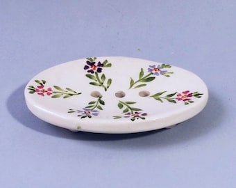 May include: White ceramic soap dish with a floral design. The dish has three drainage holes and is decorated with small purple, pink, and green flowers.