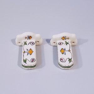 May include: Two white ceramic towel bars with green, yellow, and blue floral designs. The bars have a decorative, ornate design.