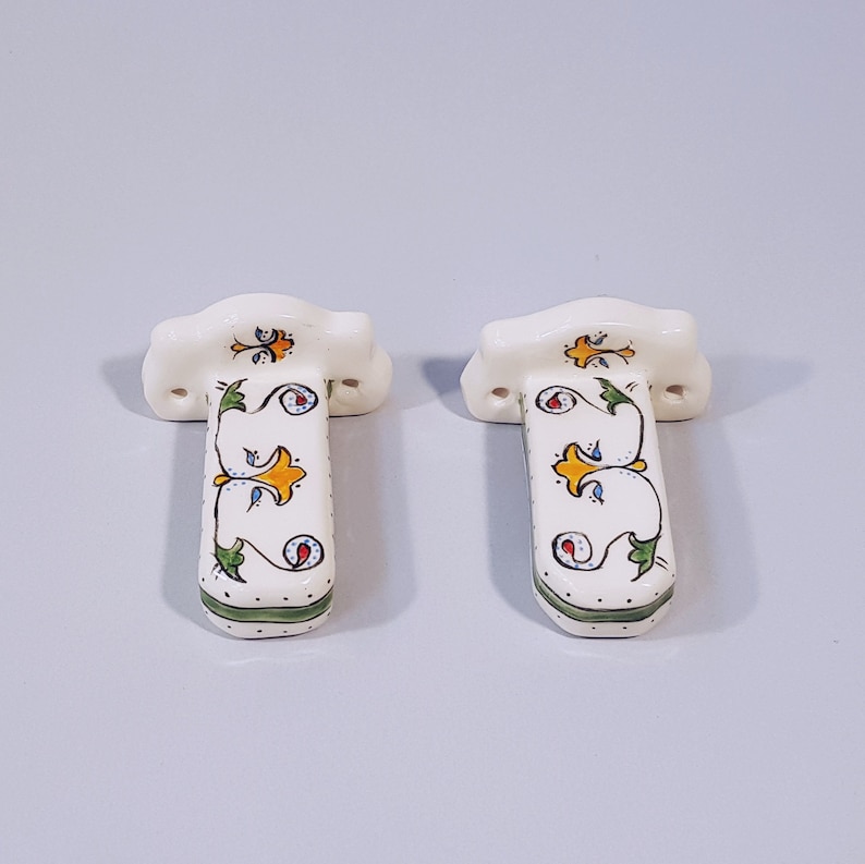 May include: Two white ceramic towel bars with green, yellow, and blue floral designs. The bars have a decorative, ornate design.