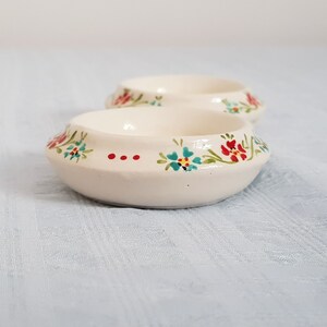 May include: Two small white ceramic bowls with a floral design. The bowls have a raised rim and are decorated with red, blue, and green flowers.