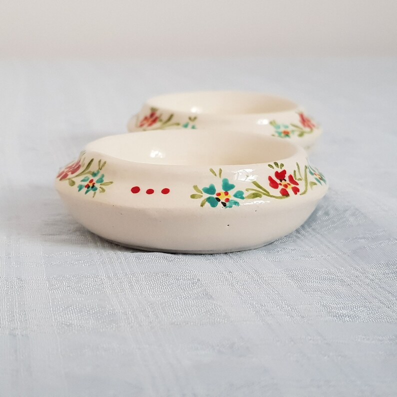 May include: Two small white ceramic bowls with a floral design. The bowls have a raised rim and are decorated with red, blue, and green flowers.
