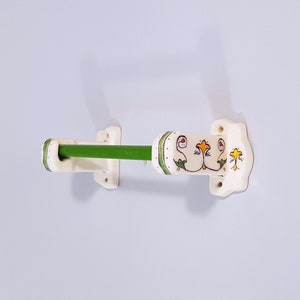 May include: White ceramic towel rack with green metal bar and floral design. The rack has a decorative design with green leaves and yellow and red flowers.