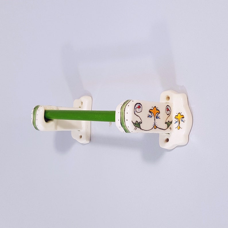 May include: White ceramic towel rack with green metal bar and floral design. The rack has a decorative design with green leaves and yellow and red flowers.