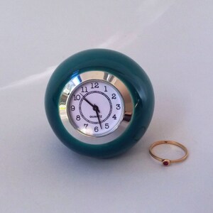 May include: A teal green ceramic clock with a white face and silver trim. The clock is round and has a ring with a red stone.