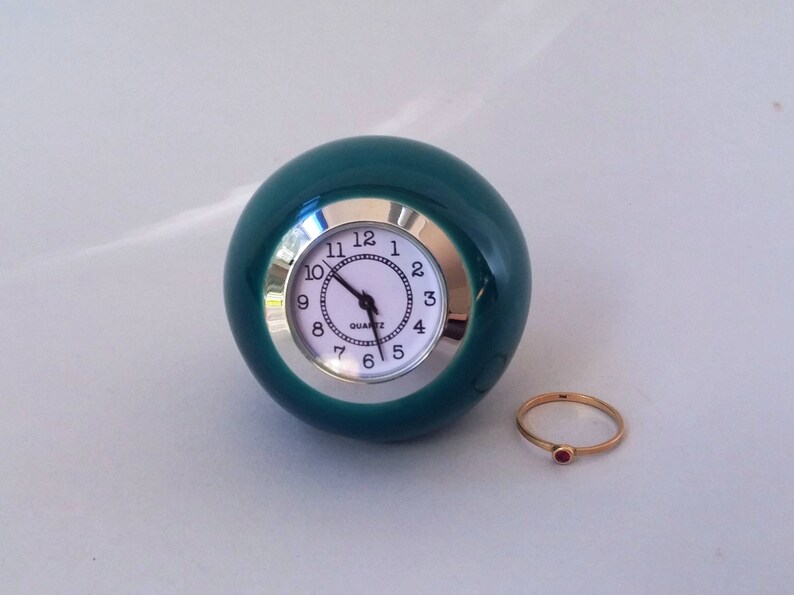 May include: A teal green ceramic clock with a white face and silver trim. The clock is round and has a ring with a red stone.