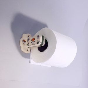 May include: White ceramic paper towel holder with a floral design in green, orange, and blue. The holder is mounted to a wall and has a roll of white paper towels on it.