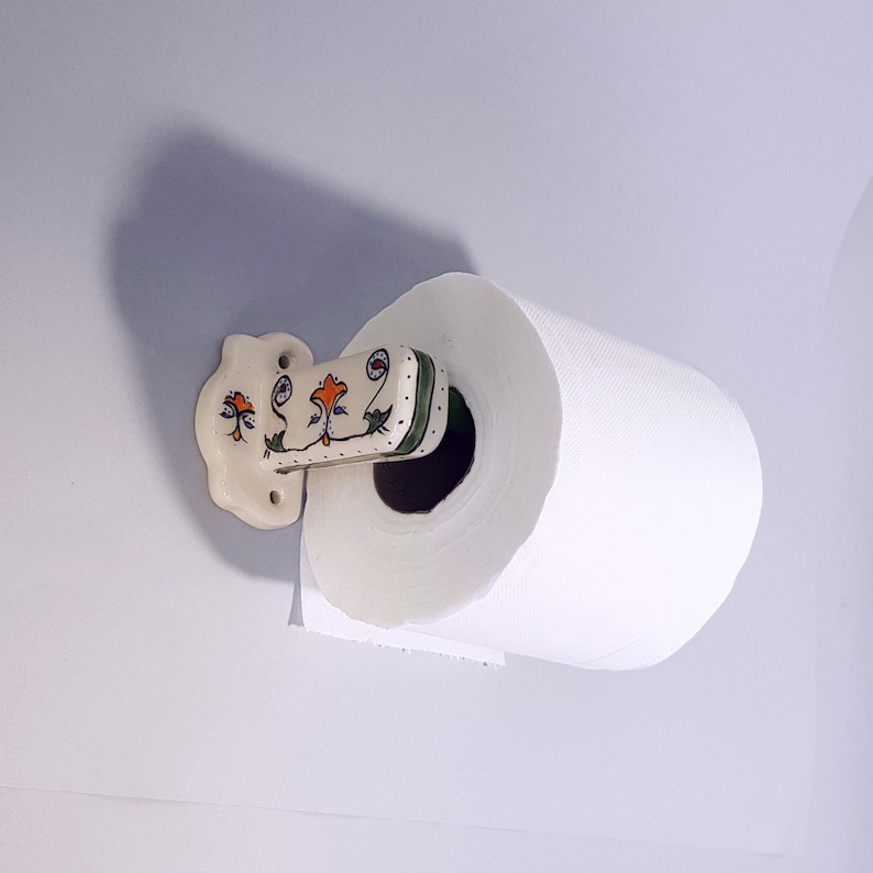 May include: White ceramic paper towel holder with a floral design in green, orange, and blue. The holder is mounted to a wall and has a roll of white paper towels on it.