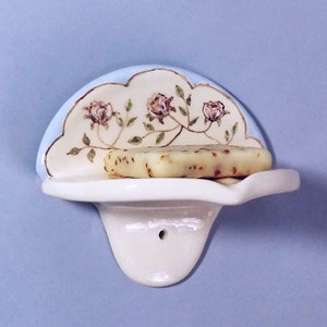 May include: A white ceramic soap dish with a scalloped edge and a floral design. The dish has a small hole on the back for hanging. A bar of soap is sitting on the dish.