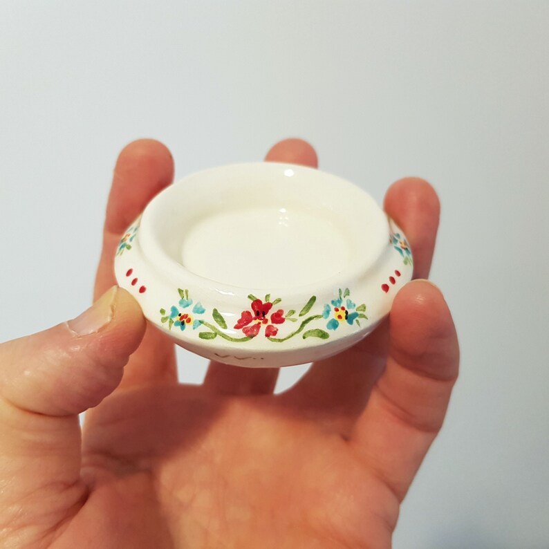 May include: A small white ceramic dish with a floral design. The dish has a raised rim and a flat bottom. The floral design features red, blue, and green flowers.
