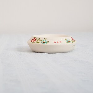 May include: A small white ceramic bowl with a floral design. The bowl has a scalloped edge and features red, green, and blue flowers.