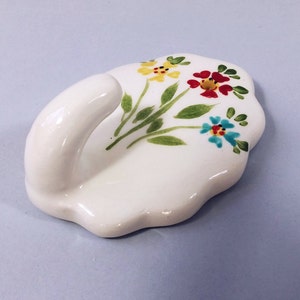 Könnte beinhalten: Weißer Keramik-Wandhaken in Blattform mit bemaltem Blumendesign. Die Blumen sind gelb, rot und blau.