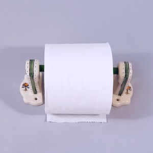 May include: White ceramic toilet paper holder with green and yellow accents. The holder has a roll of white toilet paper on it.