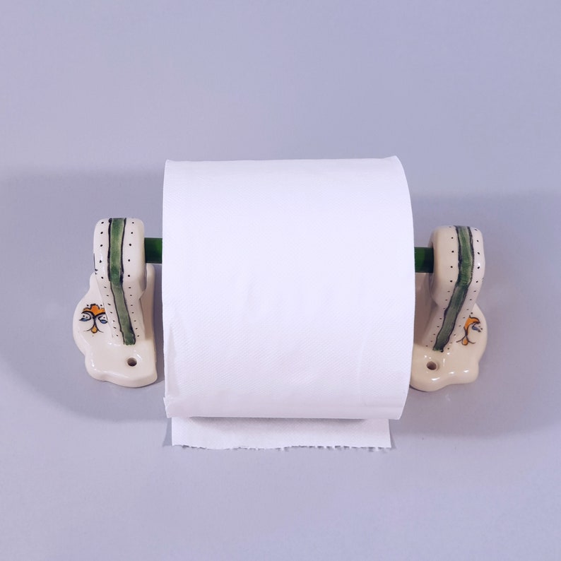 May include: White ceramic toilet paper holder with green and yellow accents. The holder has a roll of white toilet paper on it.