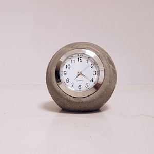 May include: A round, gray concrete clock with a silver face and black hands. The clock face has black numbers and the word "QUARTZ" printed on it.