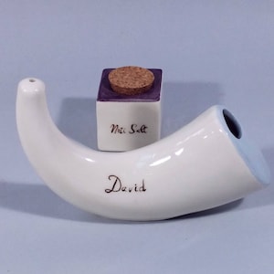 Op de afbeelding: Witte keramische hoorn met het woord "David" erop geschreven. Een kleine paars-witte keramische pot met een kurken deksel staat op de hoorn. De pot heeft de tekst "Mini Salt" op de voorkant.
