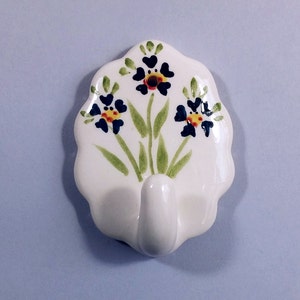 May include: White ceramic hook with a floral design. The hook features three blue, yellow, and red flowers with green leaves.