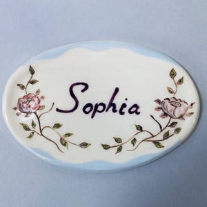 May include: Oval ceramic dish with the name "Sophia" in dark purple script. The dish is white with a light blue border and floral accents featuring pink roses and green leaves.