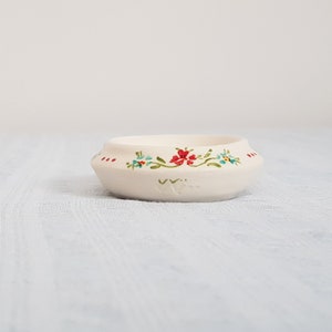 May include: A small white ceramic bowl with a floral design. The bowl has a scalloped edge and features red, blue, and green flowers.