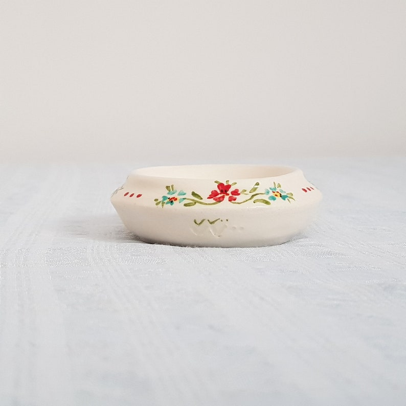 May include: A small white ceramic bowl with a floral design. The bowl has a scalloped edge and features red, blue, and green flowers.