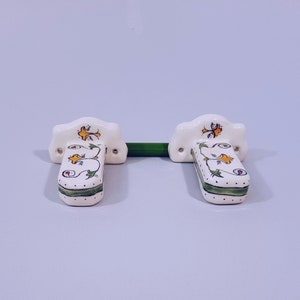 May include: A pair of white ceramic towel bar brackets with green, yellow, and blue floral designs. The brackets are mounted on a white wall.