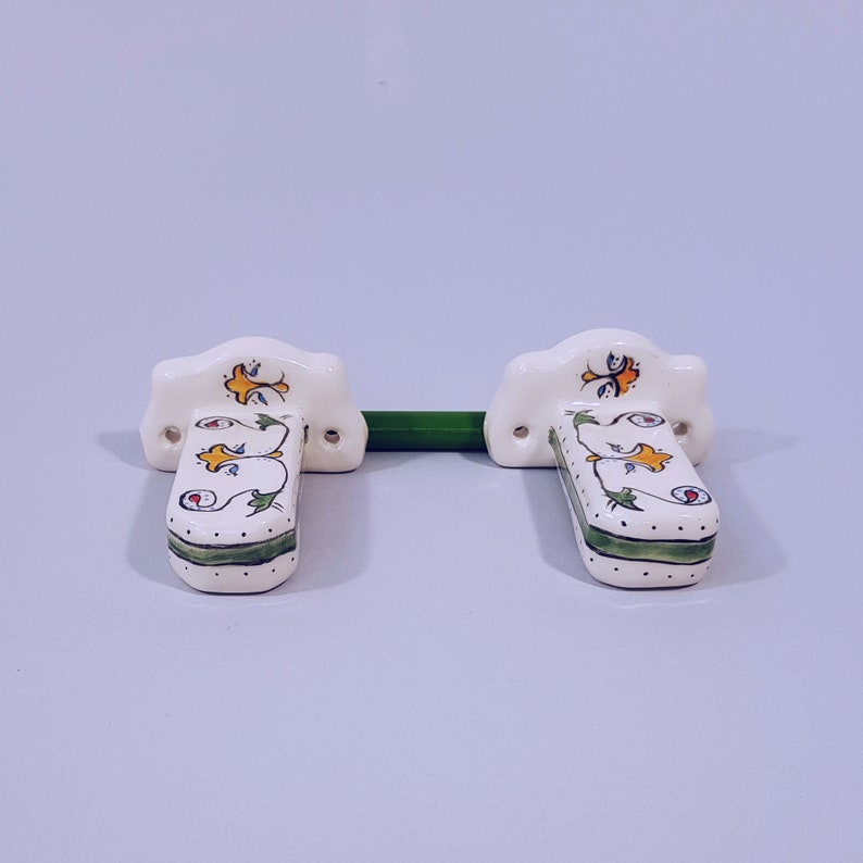 May include: A pair of white ceramic towel bar brackets with green, yellow, and blue floral designs. The brackets are mounted on a white wall.