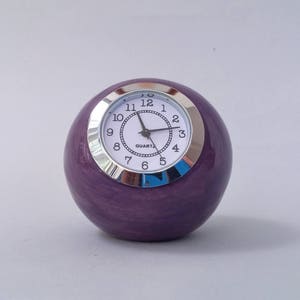 Puede incluir: Un reloj redondo morado con un borde plateado y una esfera blanca. La esfera del reloj tiene agujas negras y números negros. El reloj dice "QUARTZ" en la esfera.