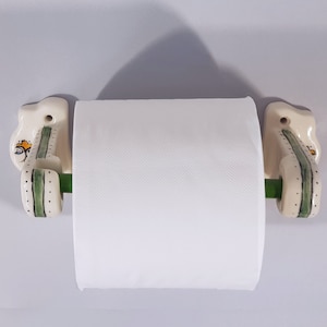 May include: White ceramic toilet paper holder with green and white floral designs. The holder has a green metal rod for the paper roll.