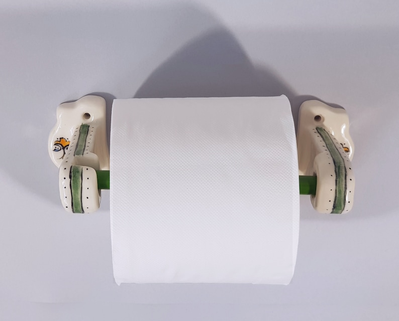 May include: White ceramic toilet paper holder with green and white floral designs. The holder has a green metal rod for the paper roll.