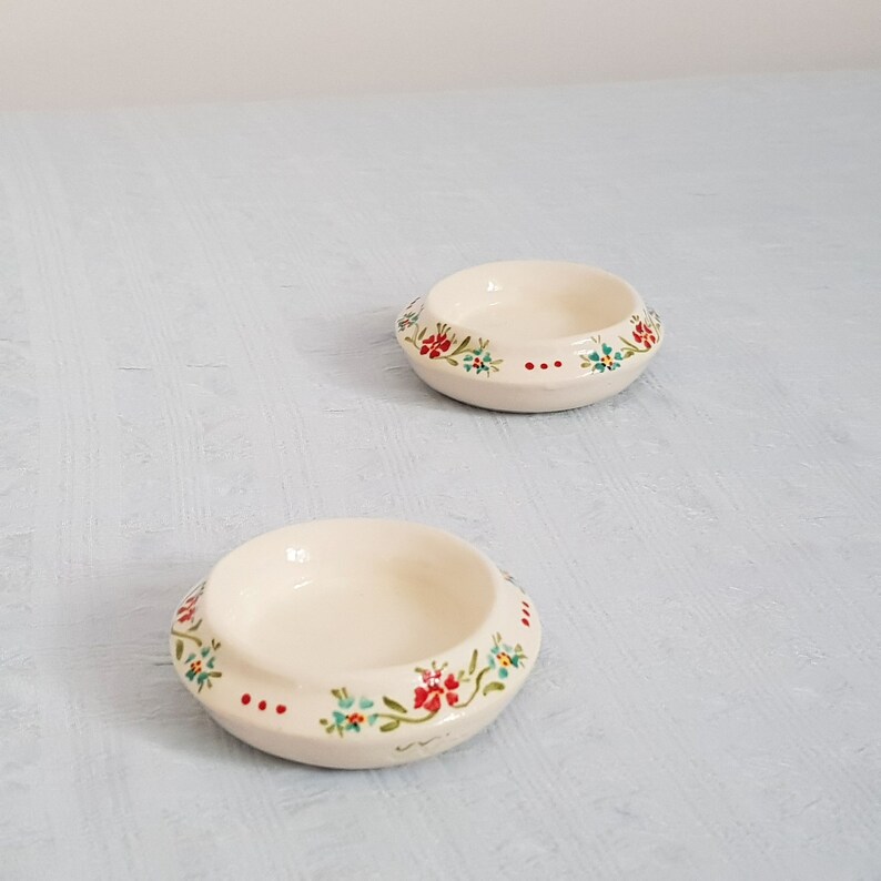 May include: Two small white ceramic bowls with a floral design. The bowls have a raised rim and a floral pattern in red, blue, and green.